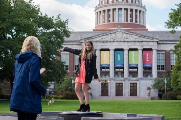 A young adult poses for a photo in front of the University of Rochester's Rush Rhees Library.