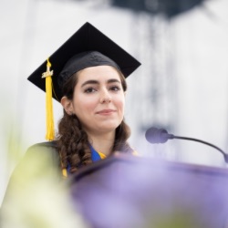 A young woman with long, dark hair wears a black graduation cap with a yellow tassel 和 gown. She st和s at a podium with a microphone, appearing to give a speech. The background is blurred, emphasizing her as the focal point.