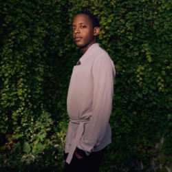 A person with short hair stands outdoors against a lush green wall of foliage. 他们穿着浅色的衣服, long-sleeve shirt and are looking slightly to the side with a calm expression. The lighting casts a soft shadow, creating a warm, natural ambiance.